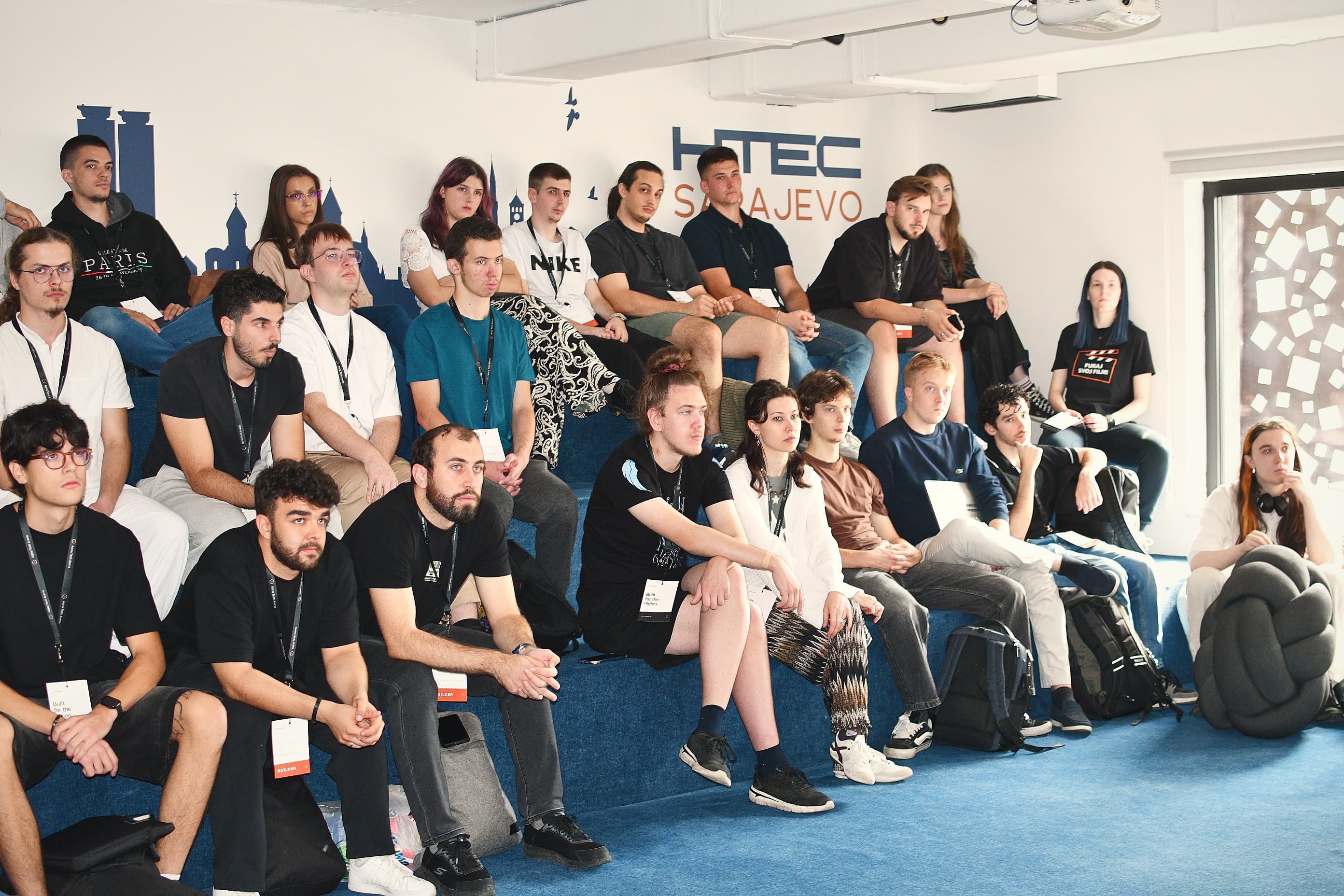 Builders seated on the tiered blue seating at HTEC Sarajevo, listening during the opening of Adria Hack Sarajevo.